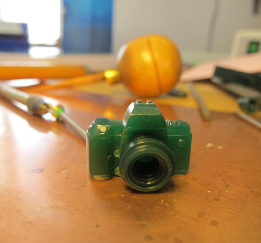 Petite sculpture verte en forme d'appareil photo sur une surface en bois, avec des outils flous en arrière-plan.