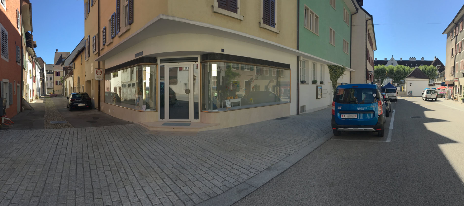 Rue pavée avec des bâtiments de chaque côté, une voiture bleue garée à droite et une vitrine d'angle à gauche.