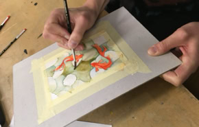Personne peignant une aquarelle sur une feuille de papier fixée avec du ruban adhésif, pinceaux à proximité.