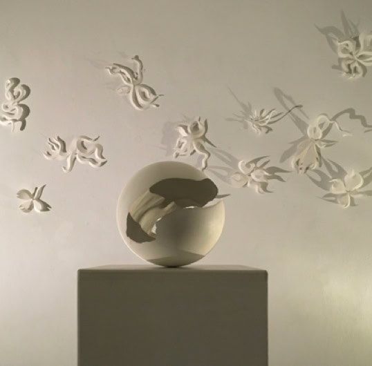 Sculpture en forme de sphère sur un socle, entourée de papillons blancs en relief sur un mur.
