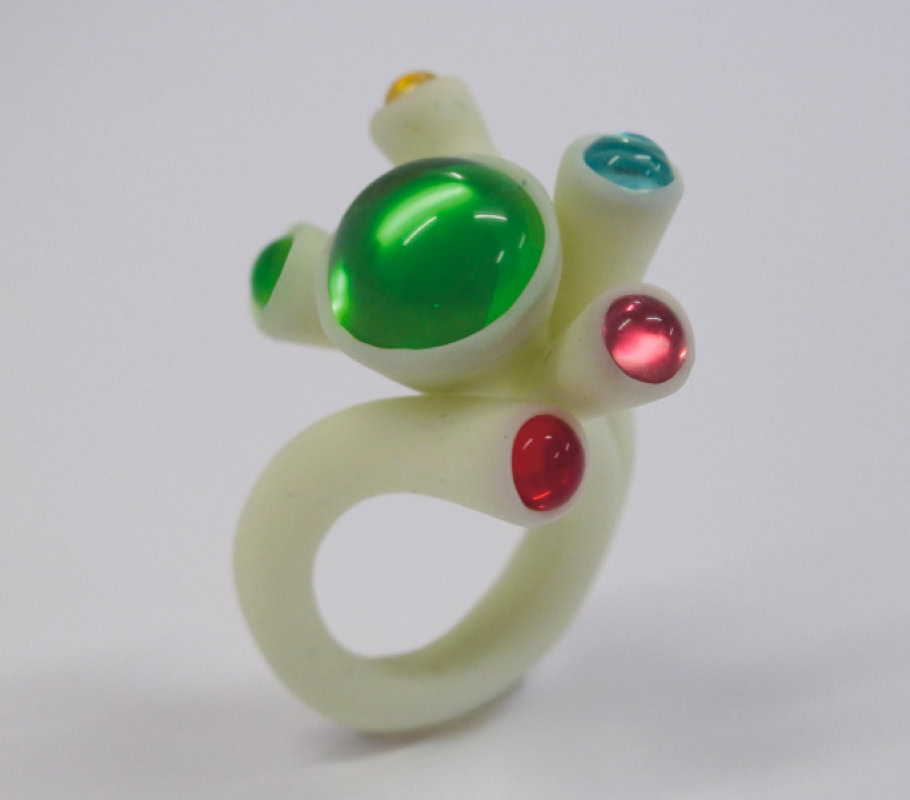Bague blanche avec des pierres vertes, rouges et bleues sur un fond blanc.