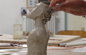 Une main sculpte une tête en argile sur une table, avec des outils et des pots en arrière-plan.
