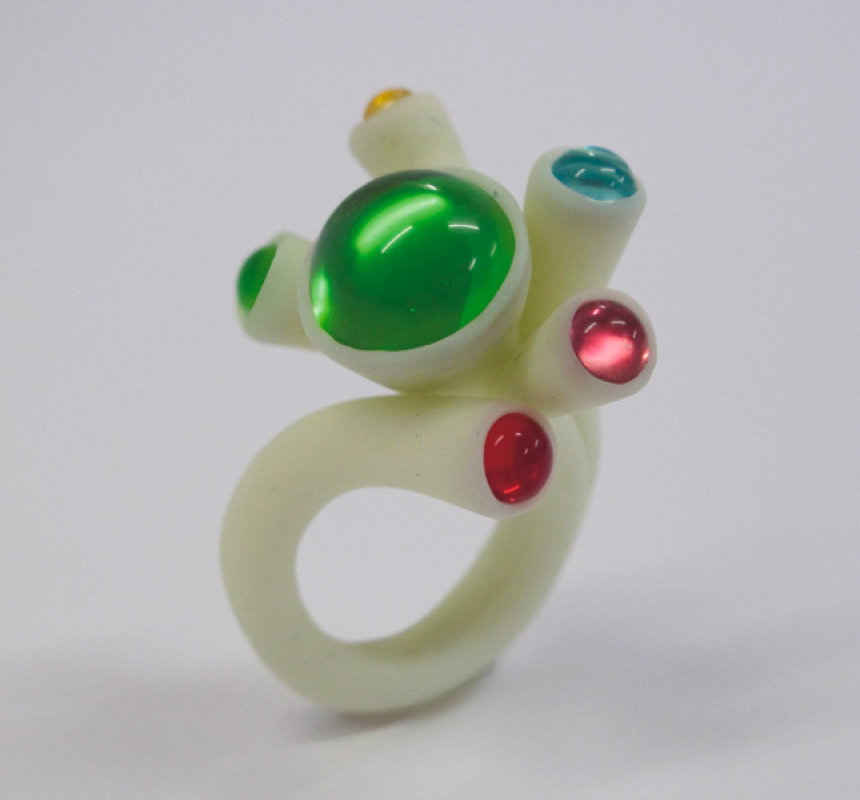 Bague blanche avec des pierres vertes, rouges et bleues sur un fond blanc.